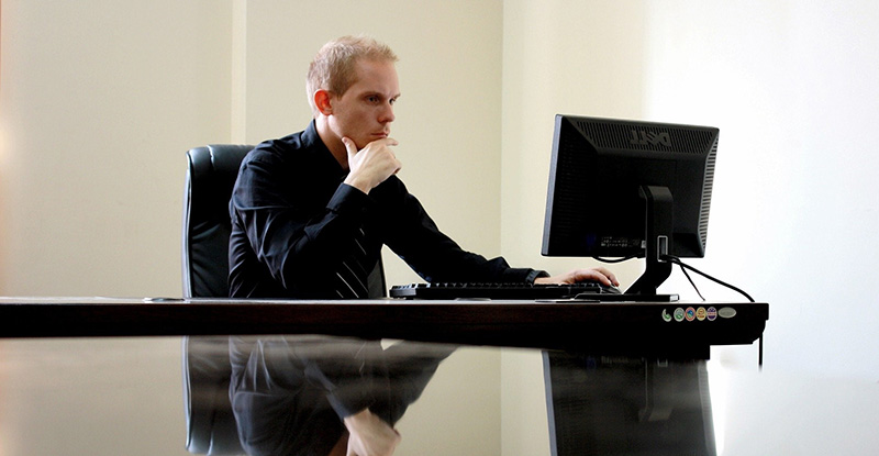 Good Character when applying to CPAWSB A photo of a man with his hand resting under his chin and looking at his computer. His reflection can be seen on the glass table.