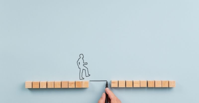 What is the Gap Verification process? A photo featuring a blue background with wooden blocks arranged in two straight lines, leaving a gap between them. A stick figure of a person is crossing the gap using a drawn line, created by a hand holding a marker.