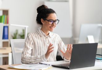A Caucasian woman smiling while video chatting on her laptop. In the background, a bookshelf and plants are visible.