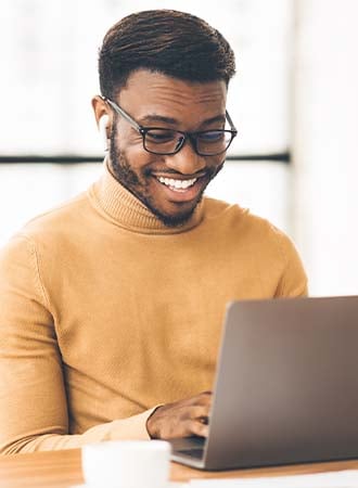 An African man wearing a mustard turtleneck and glasses, smiling while working on his laptop.
