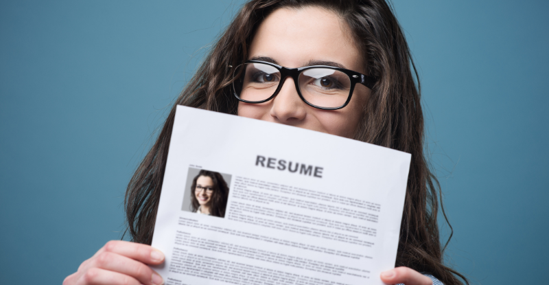 Crafting a resume for transcript assessment A photo of a woman wearing glasses holding up her resume with her picture on it. The photo is taken against a blue background.