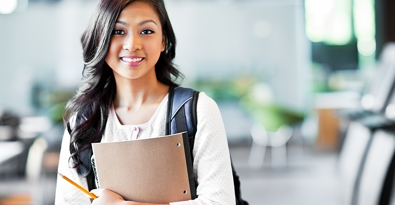 A photo of a South Asian female student smiling at the camera while carrying a backpack and holding a notebook in her hands.