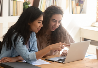 Two female students pointing at a laptop and discussing something, with bookshelves visible in the background.