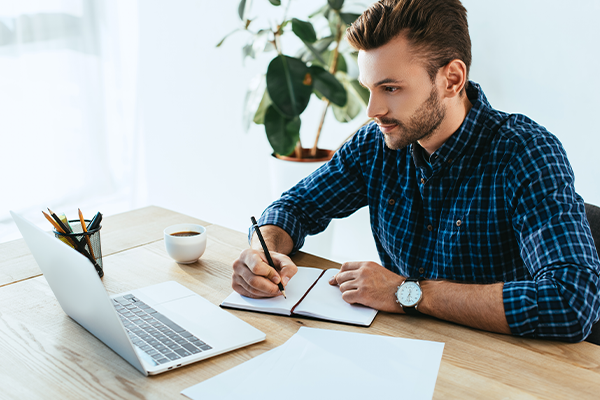 A Caucasian man wearing a blue and black checked shirt, writing in a notebook while looking at his laptop.