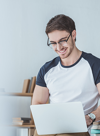 An Caucasian man wearing a t-shirt and glasses, smiling while working on his laptop.